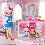 A doll playset featuring a large fashion doll with long white hair, a white cat-ear hat with a red bow, and a blue outfit, standing beside a pink bakery play kitchen with two smaller dolls. The set includes miniature appliances, a sink, and storage cabinets.