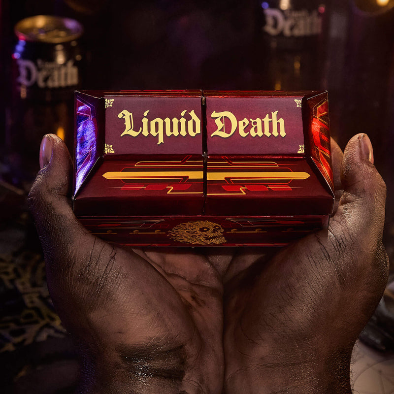 Hands that are painted black holding a Coffin like burgundy box with 'Liquid Death' in gold letters on top and a skull on  the front panel in the middle. Very dark scene behind.