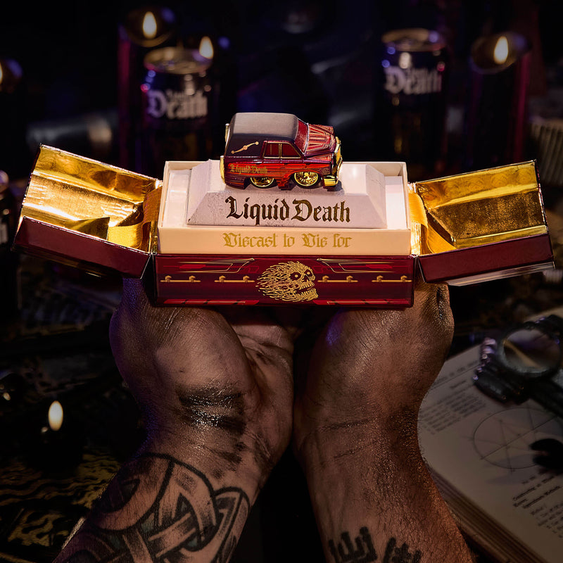 Hands that are painted black holding an open Coffin like burgundy box with 'Liquid Death' in gold letters and 'Diecast to Die For' with a skull on the front panel in the middle. Very dark scene behind.