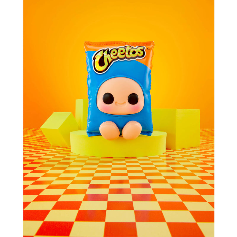 A Cheetos-themed plush toy shaped like a blue snack bag with a cartoon face, displayed on a yellow pedestal against a bright orange background with geometric blocks and a checkered floor.