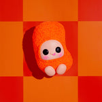 A small orange fuzzy plush toy with a pale face, large black eyes, and pink cheeks sits centered on a bold orange and red checkered background.