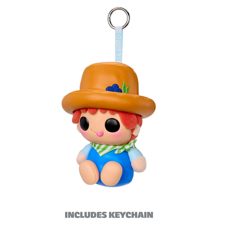 A small character keychain charm with a metal ring at the top, shown on a plain white background. The charm has a tan-orange hat, red hair, a green-and-white striped neck scarf, and blue clothing. With text 'INCLUDES KEYCHAIN'