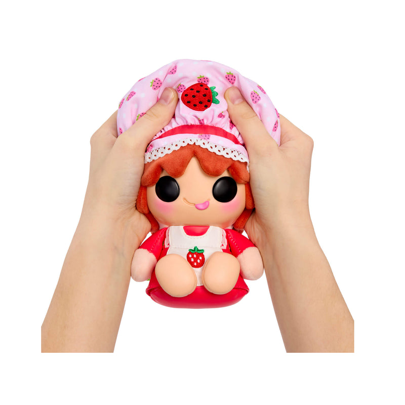 A small strawberry-themed doll being held by two hands against a plain white background. The doll has large black eyes, orange hair, a pink hat with strawberry prints, and a red-and-white outfit with a strawberry motif.