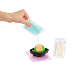 A hand holds a packet of light blue powder above a small black dish containing green and peach-colored squishy, with a pink patterned napkin underneath and another packet to the right, all on a white background.