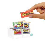 Hand holding a miniature Reese's candy bar with a display of other miniature candy packages on a white background.