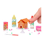 A hand putting half the roof on the house sitting on a white, round table with gumdrops. A bottle of Glaze and cream sitting near table on a white background.