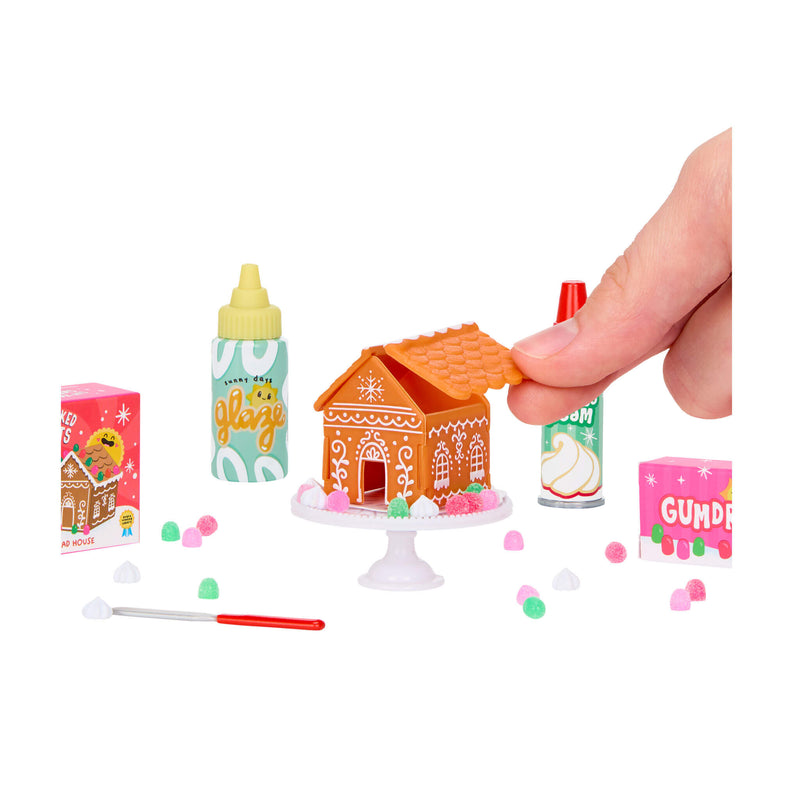 A hand putting half the roof on the house sitting on a white, round table with gumdrops. A bottle of Glaze and cream sitting near table on a white background.