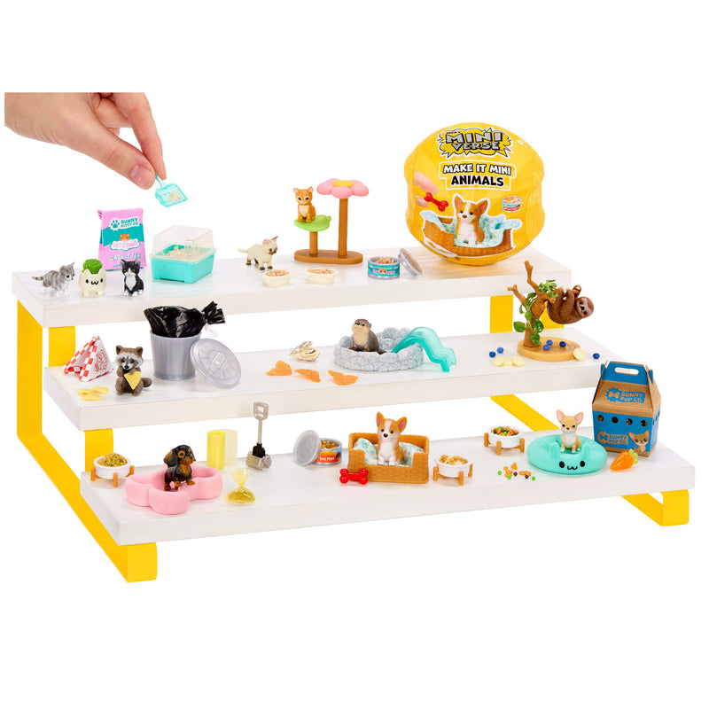 A mini animal pet display set with three white shelves and yellow legs, filled with miniature pet accessories, toys, and figurines. A human hand is placing a small item into the top slot.
