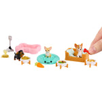 A small pet playset with miniature dog figures, bowls of food, a pink bed, a teal cushion, a basket bed, and accessories, shown on a white background with a human hand entering from the right to interact with the toy.