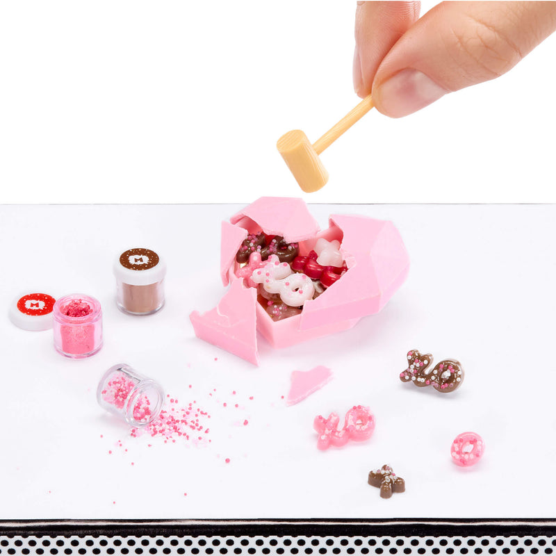 Pink heart-shaped mold being opened with a mini hammer, with decorative items on a white surface