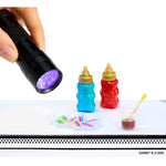 Hand holding a UV flashlight near colorful bottles and a cup with a straw on a white background.