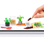 Miniature gardening set with small plants, a glass, and a hand interacting with the items on a white background.