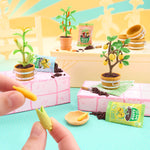 Miniature garden setup with plants and a hand holding small corn models.
