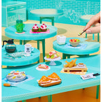 Miniature food set on a turquoise table with a hand interacting with it.