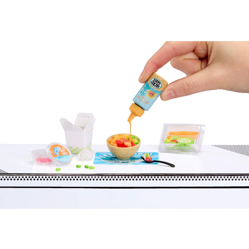 Hand pouring a small bottle of sauce onto a miniature fruit salad on a white surface.