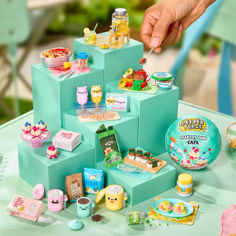 A miniature food and café set arranged on pastel blue-green display blocks, with a hand holding a spoon above the setup. The set includes tiny cups, desserts, packaged snacks, and a round ball labeled 'Mini Cafe'.