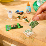 A close-up of a hand placing miniature  food items on a light wood surface, including small sushi pieces, a cup with a spoon, a bottle labeled 'Rice Vinegar,' and packaged snack pieces.