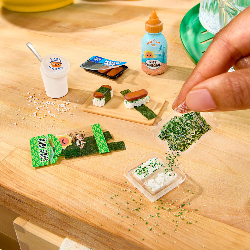 A close-up of a hand placing miniature  food items on a light wood surface, including small sushi pieces, a cup with a spoon, a bottle labeled 'Rice Vinegar,' and packaged snack pieces.