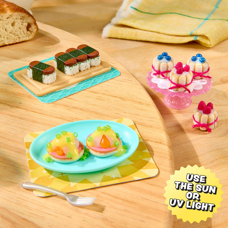 A miniature food set displayed on a wooden surface, including small sushi pieces on a brown plate, two mini eggs benedict on a blue plate sitting on a yellow patterned placemat, and three mini Charlotte cakes on a pink stand. A yellow burst in the corner reads 'USE THE SUN OR UV LIGHT'.