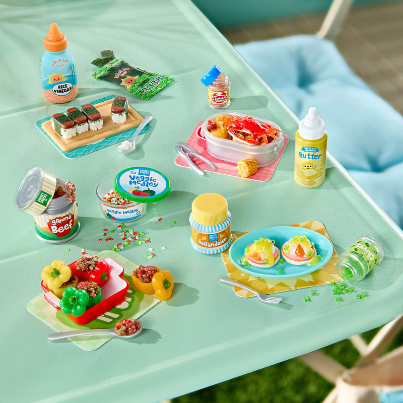 A miniature picnic scene with food items arranged on a mint green table. Visible items include a bottle labeled 'Butter', a jar labeled 'Hollandaise', a  plate with Stuffed Peppers, Sushi, Veggie pieces, and small toy utensils, with a blue chair and outdoor setting in the background.