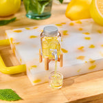 A clear glass jar with a silver cap and small wooden stand, filled with yellow liquid, has a small spout pouring the liquid into a small container. The setup is placed on a white marble tray with yellow flecks and a yellow handle, on a wooden surface with lemons and mint leaves in the background.