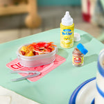 A miniature food set on a mint green tray table, featuring a plastic lunchbox filled with a red crab, a yellow bottle labeled 'Butter', a small jar with a blue lid, and silver tongs.