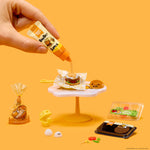 Hand pouring sauce onto a miniature burger on a yellow background
