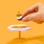 Hand holding a small burger-shaped toy with a face on an orange background