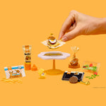 Miniature food items and a hand interacting with them on an orange background