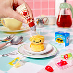 Hand squeezing a tube of "syrup" onto pancakes on a colorful tablecloth with various items around.