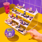 Miniature Taco Bell food set on a clear stand with a hand interacting with it, against a purple tiled wall.
