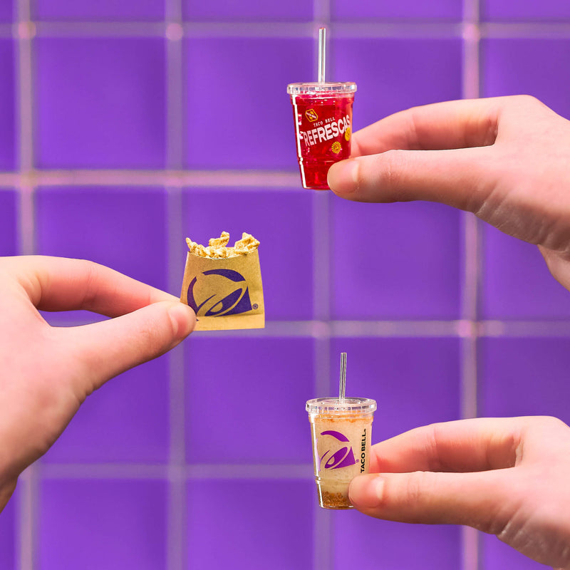 Miniature Taco Bell drinks and a bag held by hands against a purple tiled background