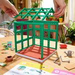 Green toy greenhouse being assembled on a table with gardening tools and plants in the background.