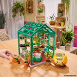 Toy greenhouse set on a table with plants and a hand interacting with it, in a room with plants in the background.