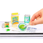 Miniature soup, pasta and packaging on a white surface with a hand interacting with them.