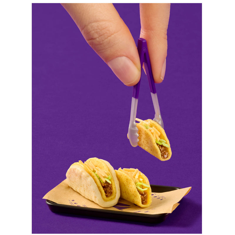 Hand holding a purple taco holder with a taco, with more tacos on a black plate below on a purple background