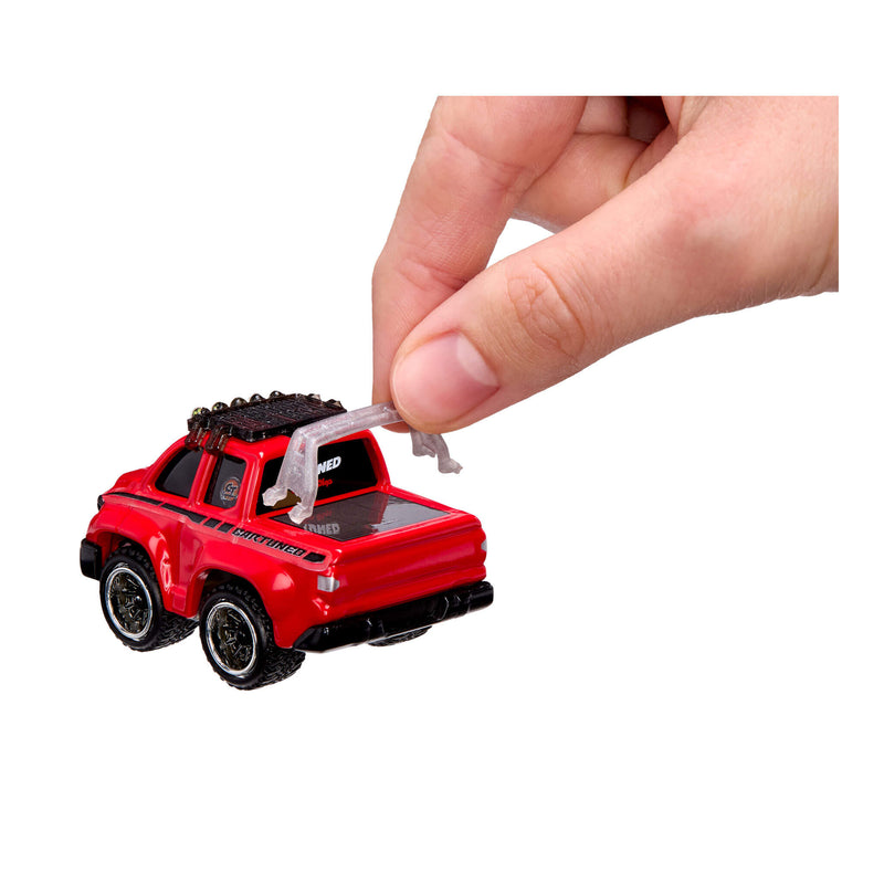 A small red toy pickup truck with black wheels and roof details, shown on a white background with a human hand holding a clear plastic strap above the bed.
