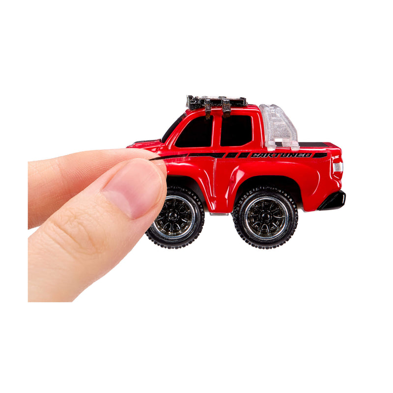 A miniature red toy pickup truck with black wheels and roof rack, a decal being placed on by a person's fingers against a plain white background.
