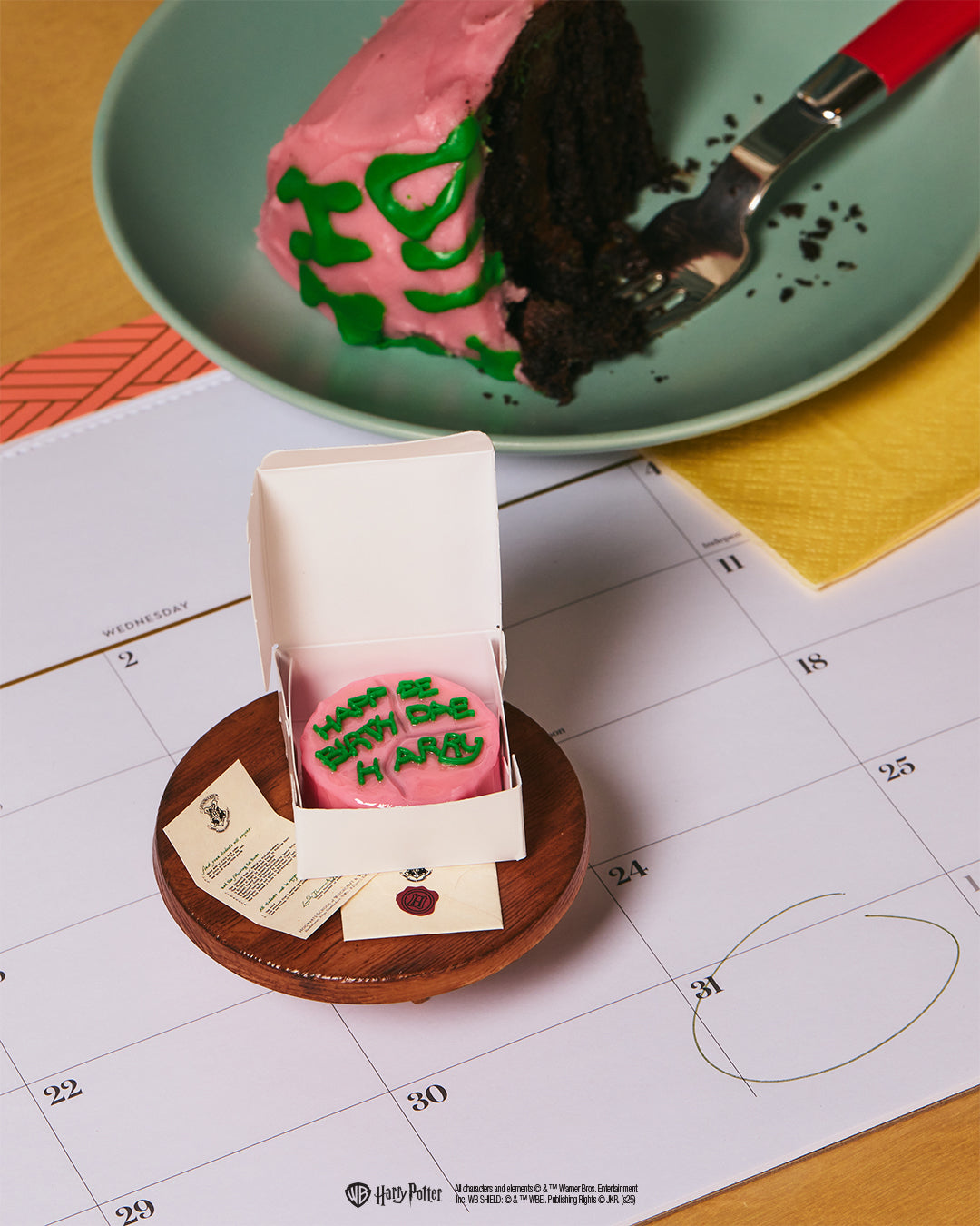 Small pink cake with green icing on a wooden stand, with a larger cake in the background on a calendar.