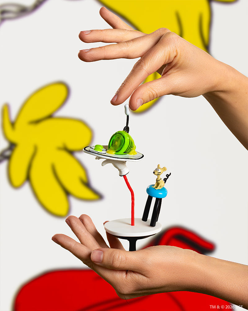 A close-up of two hands holding GREEN EGGS AND HAM BY DR. SEUSS Miniverse Special Edition against a blurred cartoon-style background with large yellow and red shapes.